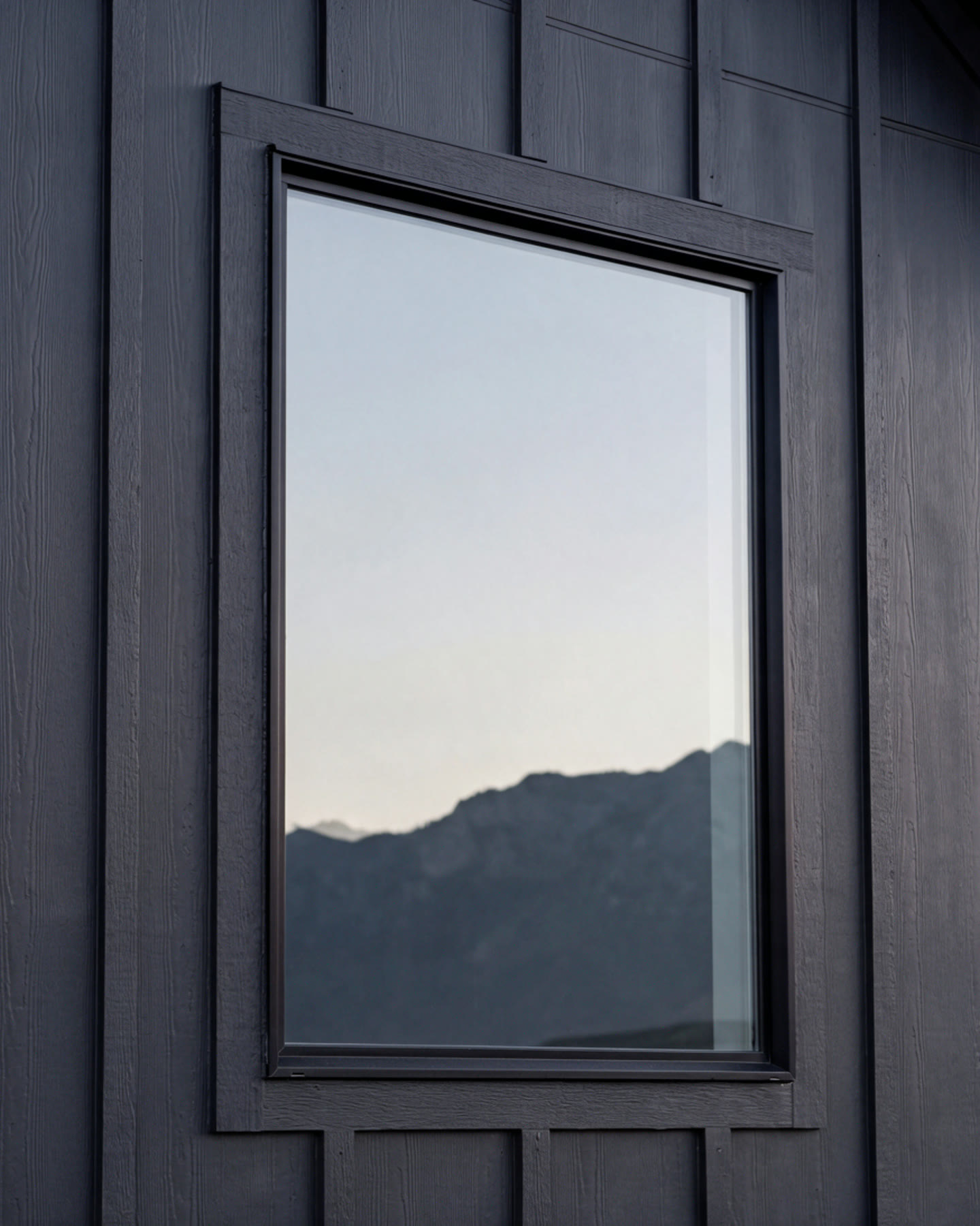 Black-framed picture window in dark charcoal board-and-batten siding, the glass reflecting a pale sky and the Wasatch ridgeline
